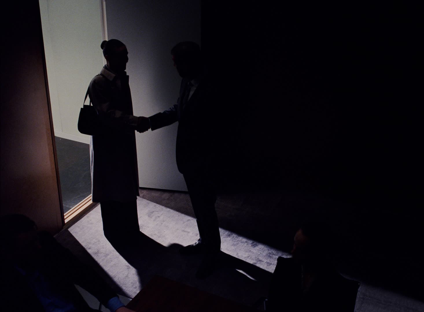 Two people shake hands in a lit doorway, seen from the darkened room next door.
