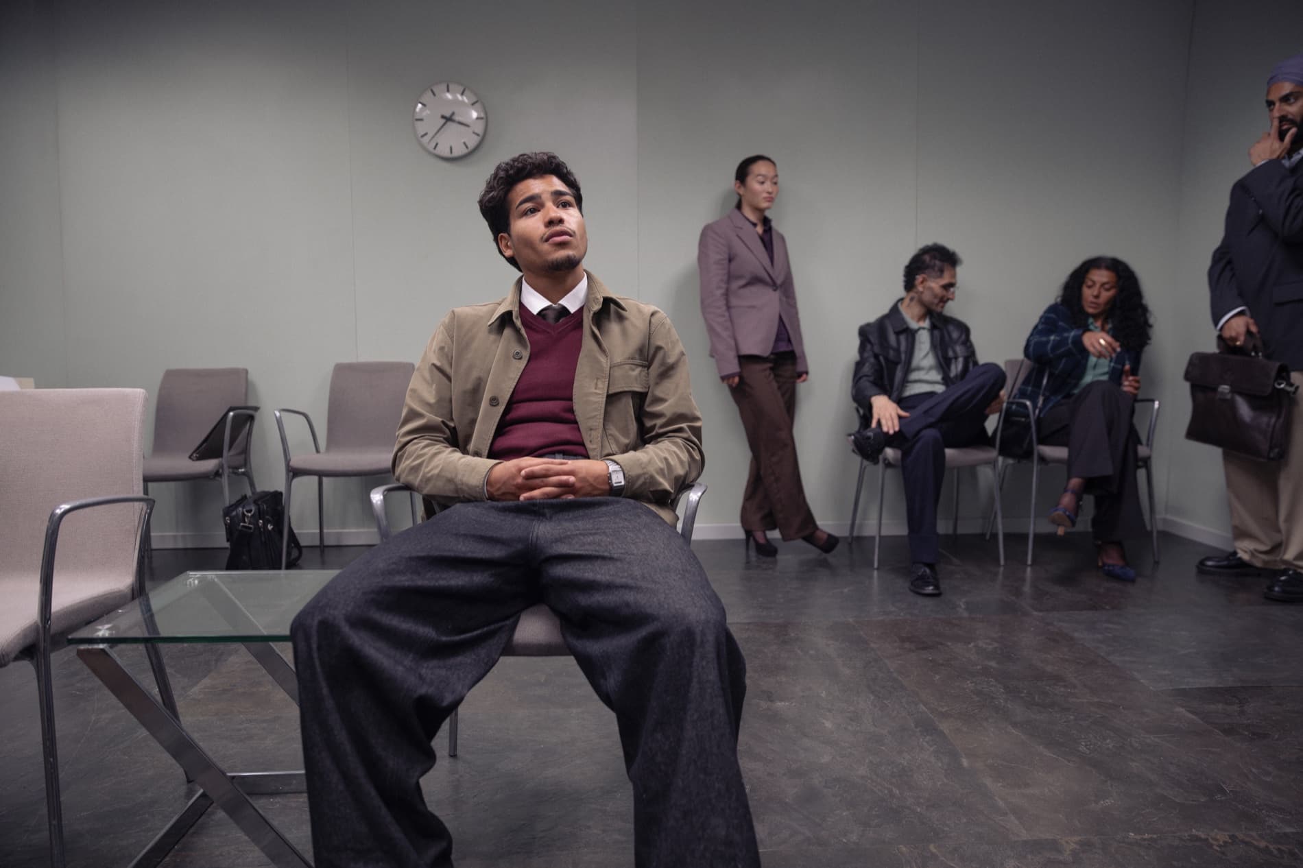 A man in a tan jacket sits waiting in a corporate room, other candidates seated and standing along the wall behind him.
