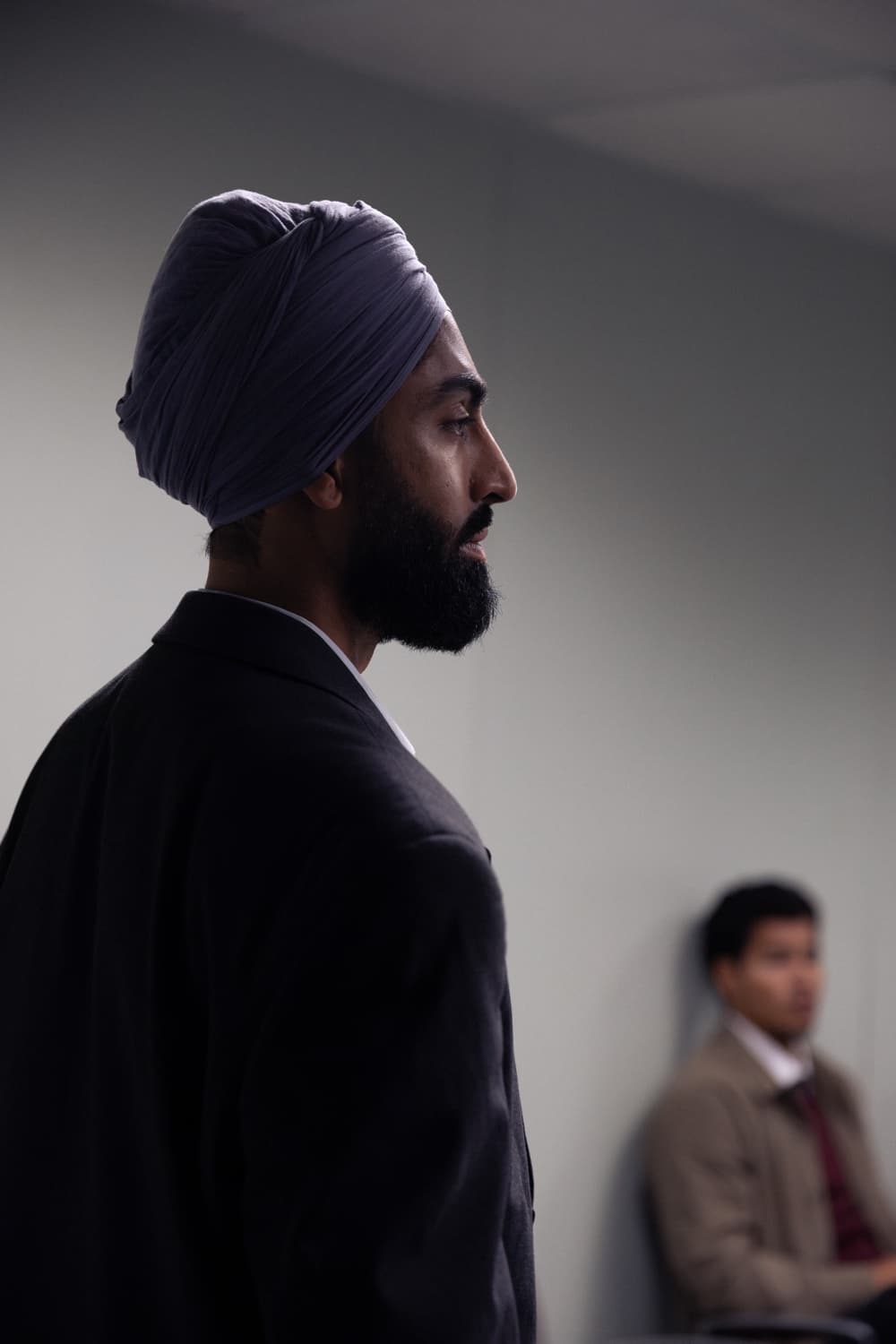 A man in a turban stands in profile against a pale wall, another candidate seated out of focus behind him.