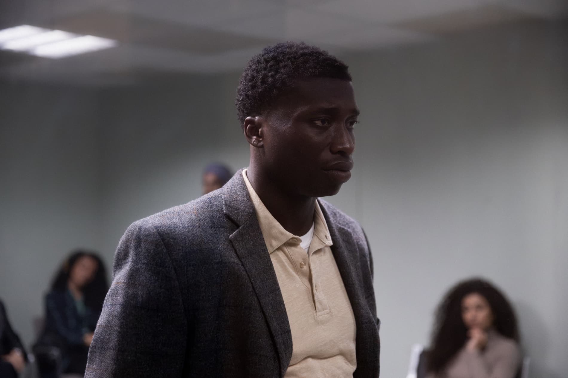 A man in a grey suit stands in a corporate room, other people out of focus behind him.