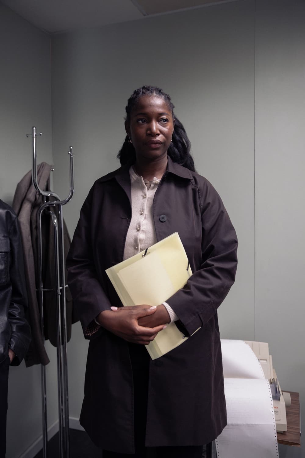 A woman in a dark coat stands holding a yellow paper folder, a coat rack beside her.
